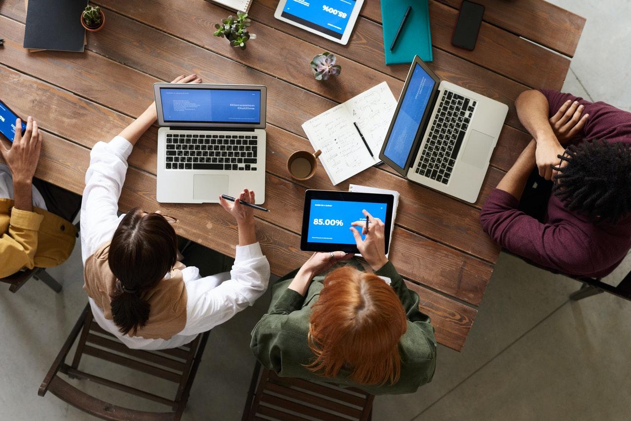 four people sitting around a table and discussing in front of their computers